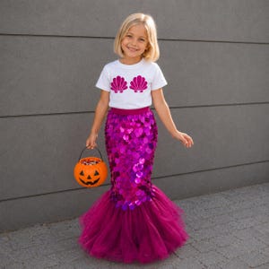 May include: A white short-sleeved t-shirt featuring two magenta seashell graphics on the chest, paired with a long, floor-length skirt covered in shiny magenta sequins and a layered tulle hem, accompanied by a small orange plastic pumpkin bucket.