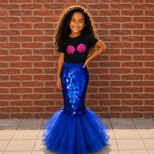 May include: A black short-sleeved t-shirt featuring two pink seashell graphics on the chest, paired with a long, fitted blue skirt covered in large, shiny circular sequins that transitions into a voluminous, layered blue tulle mermaid-style hem.