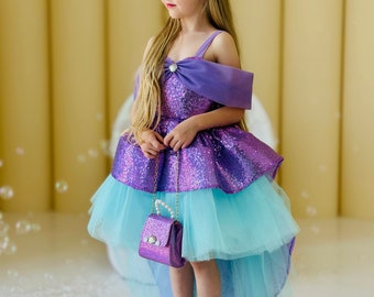 May include: A young girl wearing a purple and blue sequined dress with a high-low hemline. She is holding a small purple purse with a pearl strap. The dress has a tulle overlay and a purple sash.
