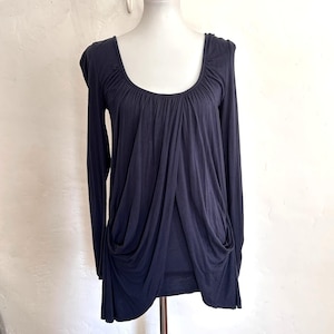 May include: A dark navy blue long-sleeve top featuring a wide scoop neckline and gathered fabric detailing at the shoulders. The front of the garment displays a draped, layered design that creates soft folds and a flowing silhouette throughout.