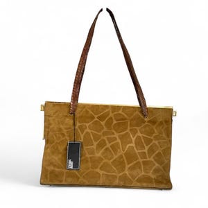 May include: A tan suede handbag with a giraffe print pattern. The bag has brown leather handles and gold-colored metal accents. A black tag hangs from the bag.