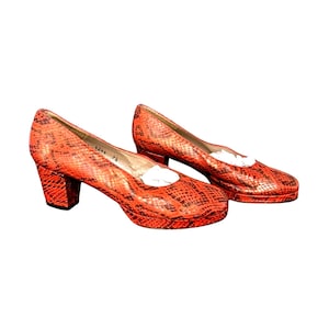 May include: A pair of orange heeled shoes with a snakeskin pattern. The shoes have a rounded toe and a block heel. The heels are approximately 3 inches high. The shoes are new, with white paper inside.