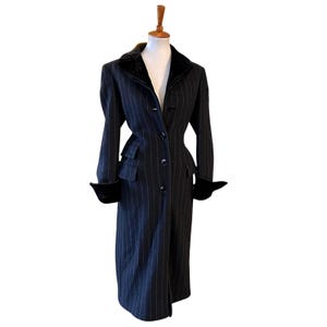 May include: A long, black pinstripe coat with a velvet collar and cuffs. The coat is buttoned up and has a tailored fit, with a small pocket on the left side. The coat is displayed on a mannequin.
