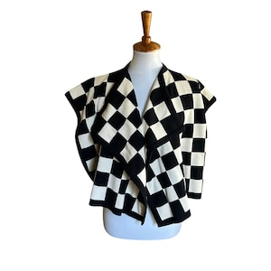 May include: A black and white checkered knit cardigan featuring an open front design with a wide draped collar and short sleeves. The garment displays a consistent geometric square pattern throughout the fabric with solid black trim.