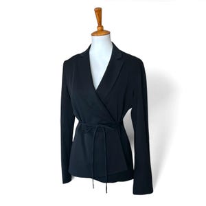 May include: A solid black blazer featuring a wrap-style front closure with a thin, matching fabric tie at the waist. The garment has long sleeves, a notched lapel collar, and a clean, structured silhouette displayed on a mannequin form.