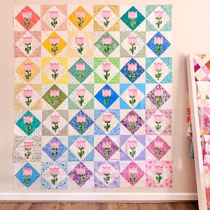 Pode incluir: Um edredom grande e colorido com um padrão repetido de aplicações de tulipas cor-de-rosa em uma grade de diamantes. O edredom apresenta uma variedade de tecidos pastel, incluindo rosa, amarelo, verde e azul. O edredom está pendurado em uma parede.