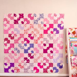 Puede incluir: Un edredón grande, hecho a mano, con un estampado floral en tonos de rosa, morado y blanco. El edredón se exhibe en una pared, mostrando su diseño de patchwork. Las flores están dispuestas en un patrón de cuadrícula, creando un textil visualmente atractivo.