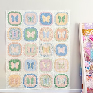 Może przedstawiać: Biała kołdra w stylu patchwork z siatką kolorowych kwadratów z motywami motyli. Każdy kwadrat ma inny wzór i kolor, w tym różowy, niebieski, zielony i żółty. Kołdra jest powieszona na białej ścianie.