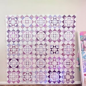 May include: A purple and white quilt with a patchwork design. The quilt is made up of squares with different patterns and is hanging on a wall.