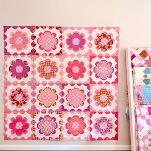 May include: A large, square quilt with a floral pattern in shades of pink, red, and orange. The quilt is made up of multiple square blocks, each featuring a flower design. The quilt is hanging on a wall, showcasing its intricate patchwork design.