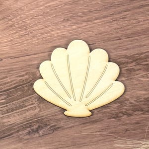 May include: A light-colored, flat wooden cutout in the shape of a scallop shell. The object features a smooth surface with engraved, thin, radiating lines that define the shell's segments, creating a simple, symmetrical fan-like silhouette.