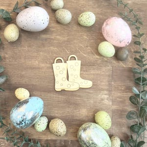 May include: Wooden cutout of rain boots with decorative eggs in various colors and patterns. The eggs are speckled, pastel, and metallic, arranged on a wooden surface with greenery. A spring or Easter theme.