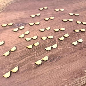 Könnte beinhalten: Eine Sammlung kleiner, flacher, goldfarbener Metallzuschnitte in Form von Brillen. Jedes Teil weist eine glatte, reflektierende Oberfläche und einen ausgeprägten Steg auf, der zwei abgerundete Brillengläser verbindet, angeordnet in einem Streumuster.