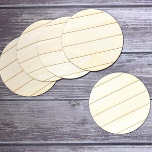 Puede incluir: Un conjunto de seis posavasos de madera sin terminar. Cada posavasos es redondo con líneas diagonales grabadas. Los posavasos son de color beige claro y están dispuestos sobre una superficie de grano de madera oscura.