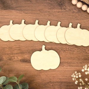May include: A set of eight flat, light-colored wooden pumpkin cutouts. Seven of the pumpkin shapes are arranged in a horizontal row, while one individual pumpkin cutout is positioned directly below the row, all featuring a smooth, unfinished surface.