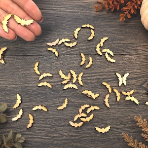 May include: A collection of small, flat, laser-cut wooden bat shapes scattered across a dark wood-grain surface. Each piece features a distinct bat silhouette with outstretched wings, rendered in a natural light brown wood tone with dark edges.