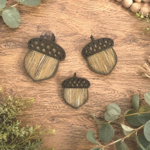 Op de afbeelding: Drie eikelvormige ornamenten met donkerbruine, lasergesneden dopjes met een ruitpatroon en lichtbruine, getextureerde lichamen met verticale nerfdetails. De ornamenten variëren in grootte, met één groot, één middelgroot en één klein exemplaar.