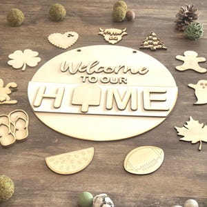 Peut inclure: Un panneau en bois avec l'inscription "Welcome to our Home", entouré de découpes en bois en forme de cœur, de trèfle, de lapin et de pastèque. Le panneau est rond et de couleur marron clair.
