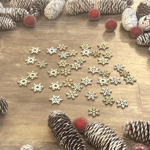 May include: A festive arrangement of pine cones, red berries, and wooden snowflakes on a wooden surface. The pine cones are brown and white, and the berries have a frosted appearance. The snowflakes are small and delicate.