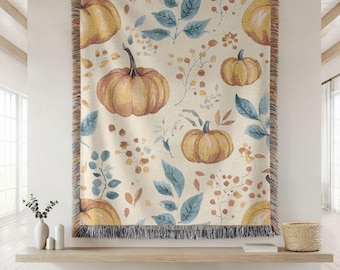 May include: A rectangular throw blanket with a cream background featuring a repeating pattern of pumpkins, blue leaves, and orange berries. The blanket has a fringed edge in shades of black, orange, and cream. The blanket is displayed on a wall.
