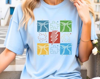 May include: A light blue t-shirt with a graphic design of a patchwork of colorful squares. Each square features a different design, including a puzzle piece, a bow, a lightning bolt, and a checkered pattern.