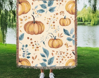 May include: A cream-colored throw blanket featuring a repeating pattern of orange pumpkins, blue-green leaves, and brown floral accents. The blanket has a fringed edge and is held up by a person wearing white sneakers.