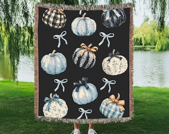 May include: A black throw blanket featuring watercolor pumpkins in various patterns and colors, including blue, white, and plaid. The pumpkins are adorned with bows. The blanket is fringed and held up by a person wearing white sneakers, with a lake and trees in the background.