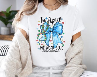 May include: White t-shirt with the text "In April We Wear Blue Autism awareness." The shirt features a large blue bow with a ribbon in the colors of the autism awareness symbol, surrounded by colorful flower accents.