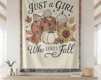 May include: Cream-colored woven throw blanket with a fall-themed design. The design includes pumpkins, a sunflower, and the text "Just a Girl Who Loves Fall". The blanket has a fringed edge and is displayed on a wooden shelf.