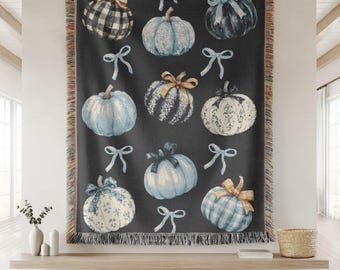 May include: A black throw blanket featuring watercolor pumpkins in various patterns and colors, including blue, white, and plaid. Each pumpkin is adorned with a bow. The blanket has a fringed edge, adding a decorative touch to the fall-themed design.