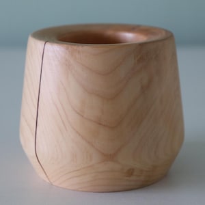 May include: A light wood vase with a unique, slightly angled design. The vase is made of natural wood and has a smooth, polished finish.