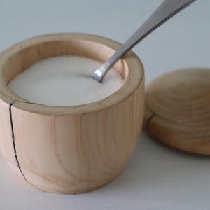 May include: A wooden salt cellar with a lid and a silver spoon inside. The cellar is filled with white salt.