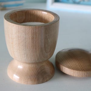 May include: A wooden container with a lid, both made of light brown wood. The container has a rounded top and a base with a flared lip. The lid is slightly domed.