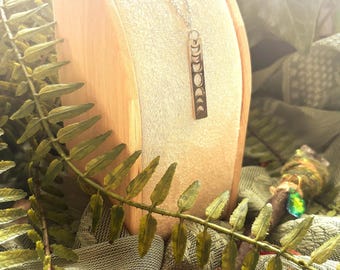May include: A silver necklace with a vertical pendant showing the phases of the moon. The necklace is displayed on a wooden stand, with green fern leaves and a pink flower in the foreground.