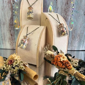 May include: Glass vial necklaces filled with dried flowers and herbs, displayed on wooden stands. The vials have cork stoppers and hang from black chains. Two small bouquets of dried flowers and rolled paper are also visible.