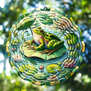 Può includere: Un mulino a vento rotondo, in stile vetrata, raffigurante una rana verde su una ninfea in uno stagno. Il design include ninfee, acqua e ninfee rosa, con un bordo verde e oro. Lo sfondo è un fogliame verde sfocato.