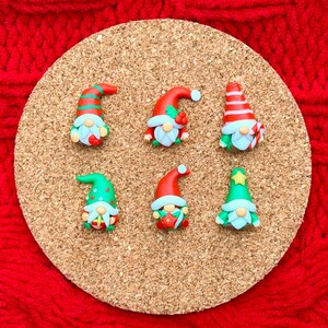 May include: Six small gnome figurines arranged on a circular cork coaster. Each gnome features a unique hat design in red, green, and white, with a light blue beard and a round nose. The gnomes display various holiday-themed accessories.