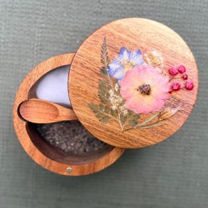 May include: A round wooden salt cellar with a matching lid featuring pressed pink and blue flowers and green foliage. The open container holds dark gray salt and includes a small wooden spoon resting inside the bowl, showcasing natural wood grain.