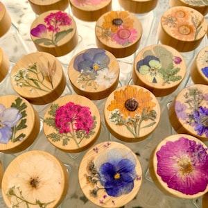 May include: A collection of circular wooden lids, each featuring a unique arrangement of pressed dried flowers in vibrant shades of pink, blue, yellow, and purple. The natural wood grain is visible on the sides of the lids, which are arranged in a grid pattern.