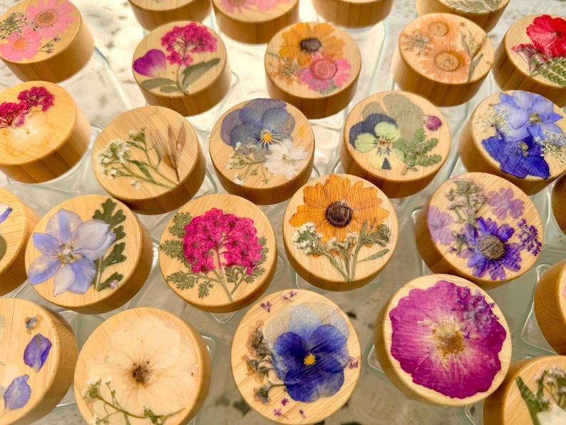 May include: A collection of circular wooden lids, each featuring a unique arrangement of pressed dried flowers in vibrant shades of pink, blue, yellow, and purple. The natural wood grain is visible on the sides of the lids, which are arranged in a grid pattern.
