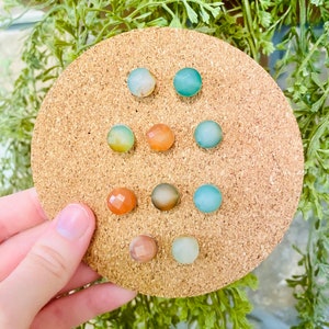 May include: A corkboard with nine round, colorful, agate stone earrings. The earrings are in shades of green, blue, and orange.