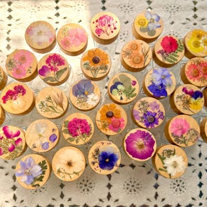 May include: A collection of circular wooden lids, each featuring a unique arrangement of pressed, dried flowers in various colors including pink, yellow, blue, purple, and white, displayed as a group of individual decorative jar toppers.