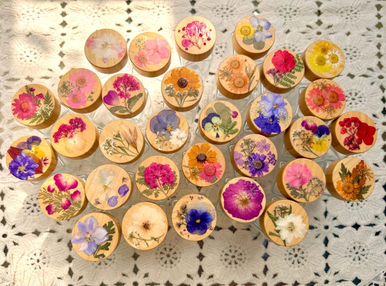 May include: A collection of circular wooden lids, each featuring a unique arrangement of pressed, dried flowers in various colors including pink, yellow, blue, purple, and white, displayed as a group of individual decorative jar toppers.