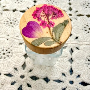 May include: A clear glass jar with a round wooden lid featuring a pressed pink hydrangea flower, a single purple petal, and green leaves embedded on the top surface. The lid has a natural light wood grain finish and a smooth, circular shape.