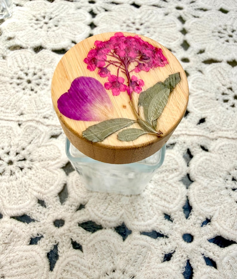 May include: A clear glass jar with a round wooden lid featuring a pressed pink hydrangea flower, a single purple petal, and green leaves embedded on the top surface. The lid has a natural light wood grain finish and a smooth, circular shape.