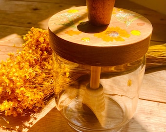 May include: A clear glass honey jar with a wooden lid decorated with yellow sunflowers and other floral designs. The jar includes a wooden honey dipper. The jar sits on a wooden surface, with dried yellow flowers in the background.