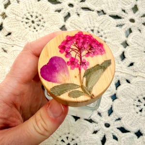 May include: A circular wooden lid for a glass container featuring a pressed pink hydrangea flower, a single dark pink petal, and green leaves embedded on the top surface. The wood grain is visible beneath the preserved botanical elements.