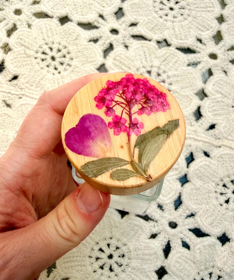 May include: A circular wooden lid for a glass container featuring a pressed pink hydrangea flower, a single dark pink petal, and green leaves embedded on the top surface. The wood grain is visible beneath the preserved botanical elements.
