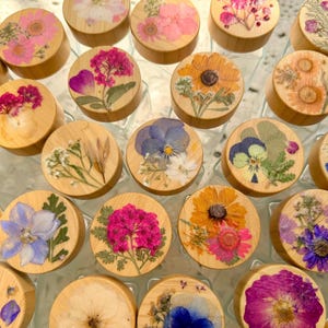 May include: A collection of circular wooden lids for jars, each featuring a unique arrangement of pressed dried flowers in vibrant shades of pink, blue, yellow, and purple, showcasing natural botanical details on a light wood surface.