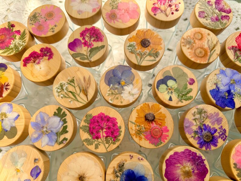 May include: A collection of circular wooden lids for jars, each featuring a unique arrangement of pressed dried flowers in vibrant shades of pink, blue, yellow, and purple, showcasing natural botanical details on a light wood surface.